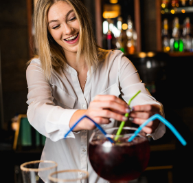 A smiling bartender at Mr MIKES Steakhouse adds colorful straws to a large shared cocktail.