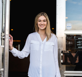 A welcoming hostess smiles while holding the door open at Mr MIKES Steakhouse, inviting guests into the restaurant.