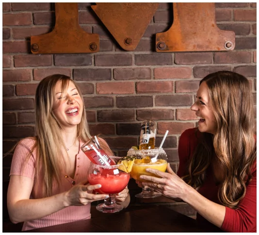 Two women share a laugh while enjoying large, colorful drinks at Mr MIKES Steakhouse, with the restaurant's rustic logo mounted on a brick wall in the background.