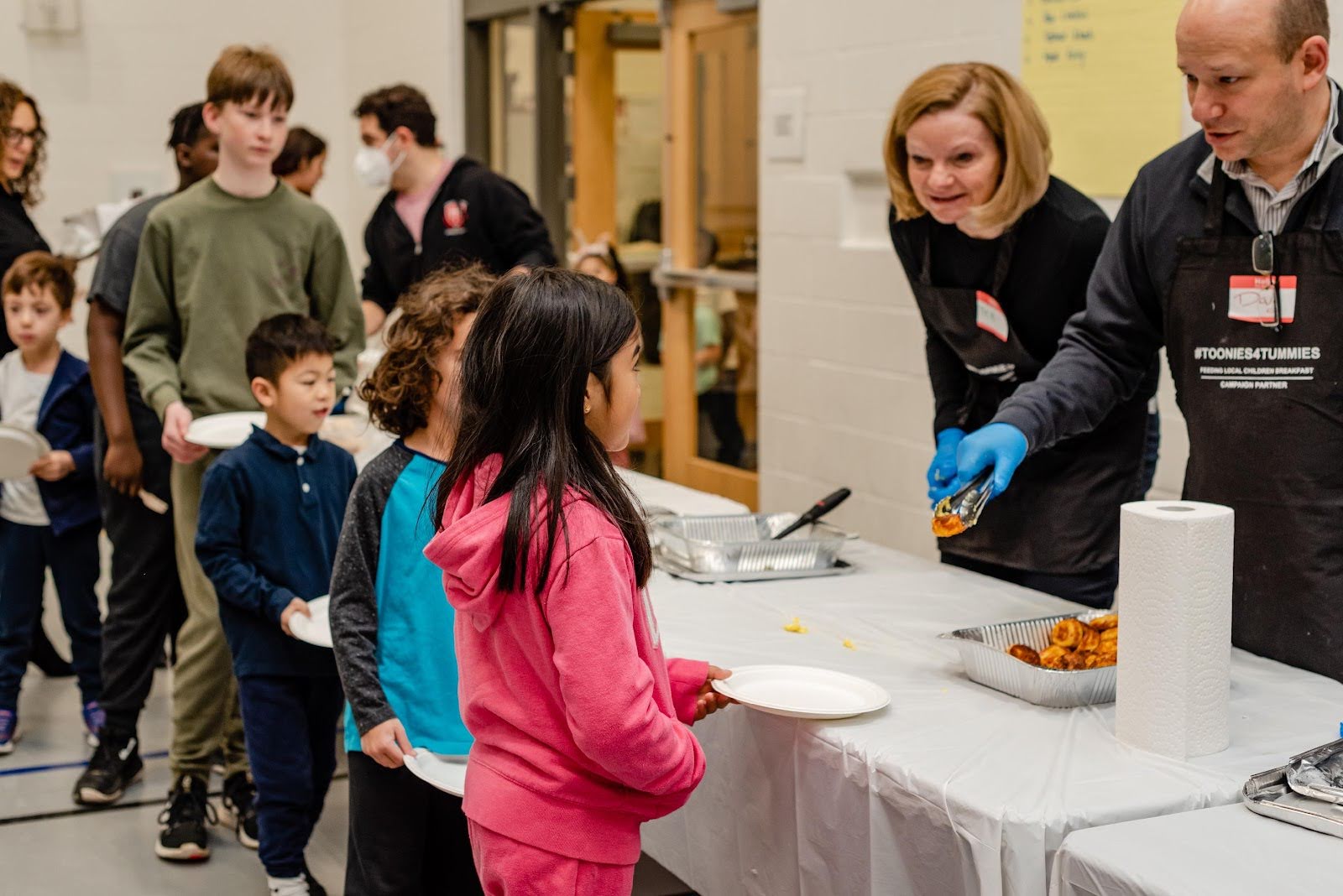 A group of children lined up indoors, receiving food served by two adults wearing gloves behind a table covered with a white cloth